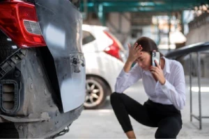 A business woman in a rental car kneels by a damaged bumper while making a phone call.